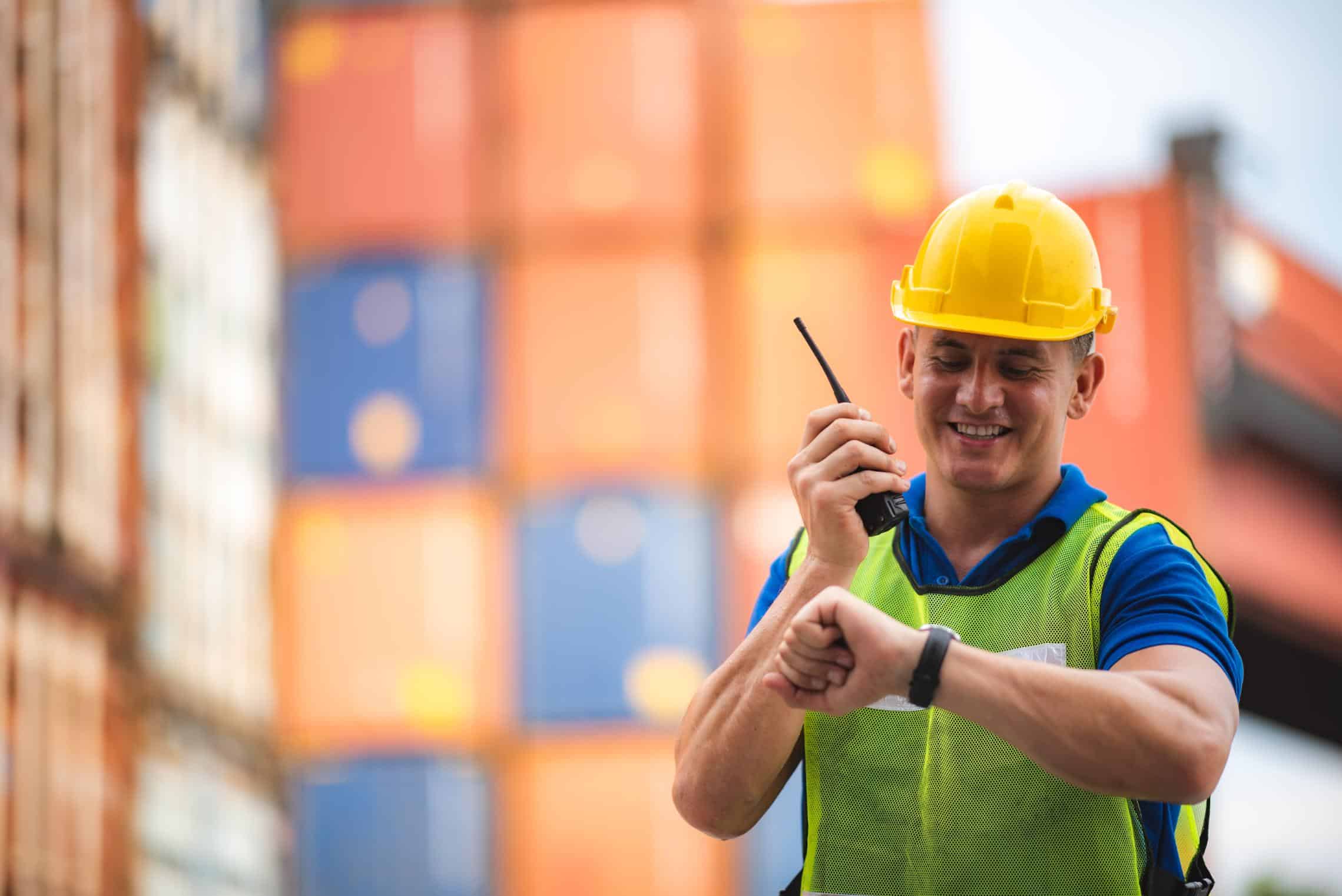 Operario con casco revisando un reloj en un entorno de construcción con contenedores de carga.